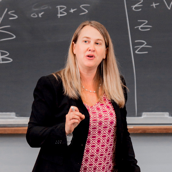 A photo of Jessica Erickson teaching. She is in front of balckboard and wearing a patterned shirt and black blazer.