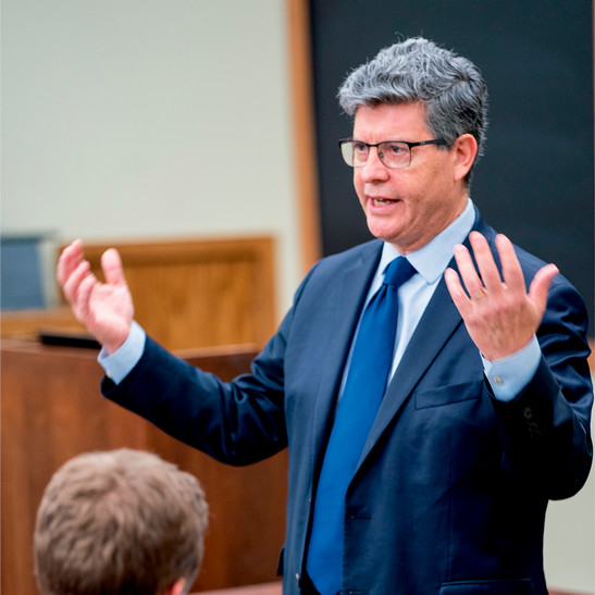 A photo of Kurt Lash teaching. He is gesturing with both hands, and he is wearing glasses and a blue suit.