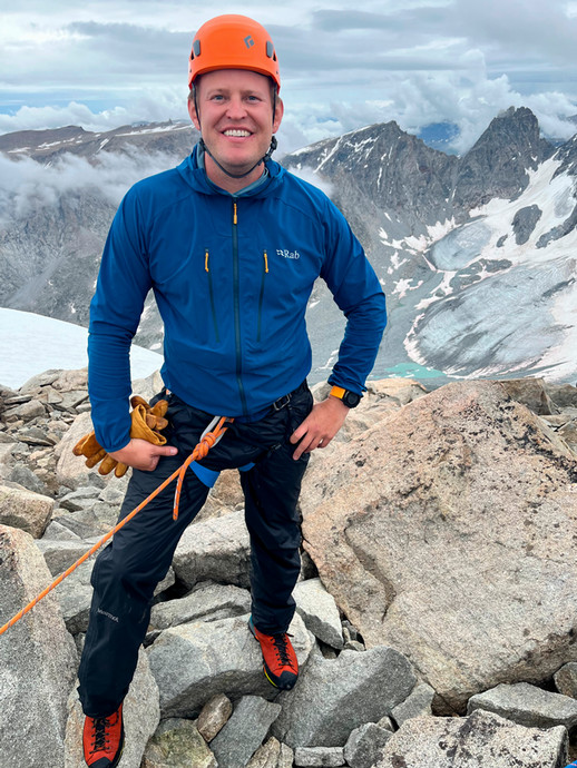 Jacob Rooksby in climbing gear atop a mountain with a view of glaciers below.