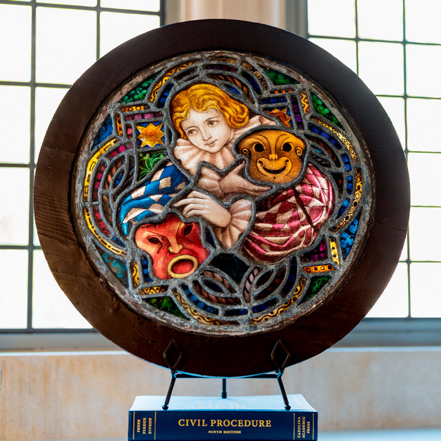 A photo of a round stained glass piece depicting a feamle figure with the masks of comedy and tragedy.