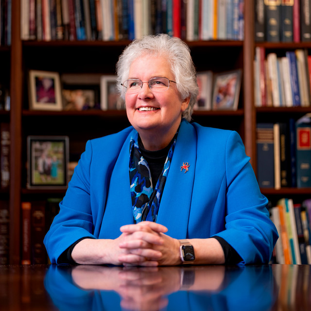 A portrait of dean Wendy Perdue. She is looking up and off camera, smiling. She is wearing a blue jacket and sitting in front of a full bookshelf.