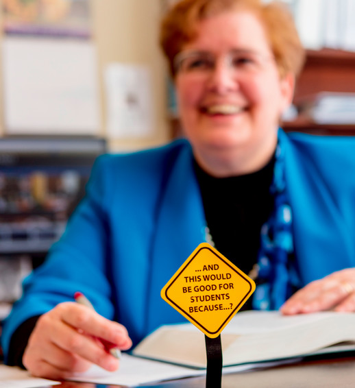 A photo of Dean Wendy Perdue at her desk. She is out of focus in the background. In focus in the foreground is a small yellow metal sign, styled like a street sign, that reads 