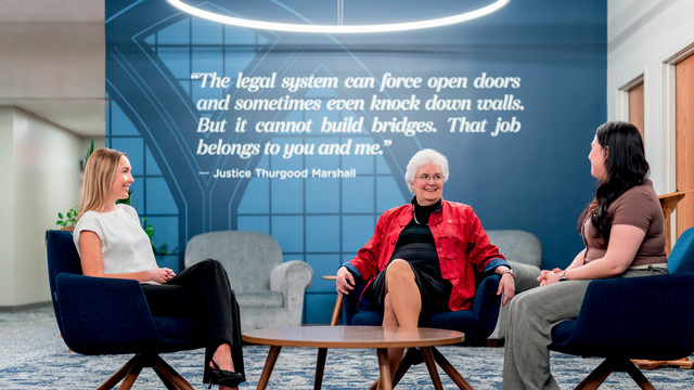 Dean Wendy Perdue, sitting and taling with two law students. They are seated in the law school commons in front of a large quote on teh wayy that reads 