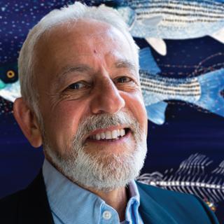A photo of Eugene Maurakis in front of one of his paintings, which depicts fish on a blue background surrounded by colorful swirls comprised of dots.