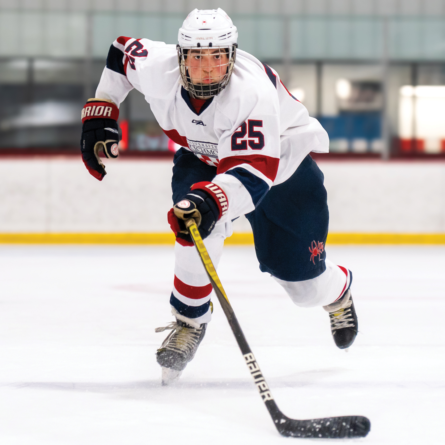 A hockey player, Ethan Gail, '25, the team captain, skates towards the camera in an indoor ice arena. He is in a University of Richmond jersey and holding a hockey stick.