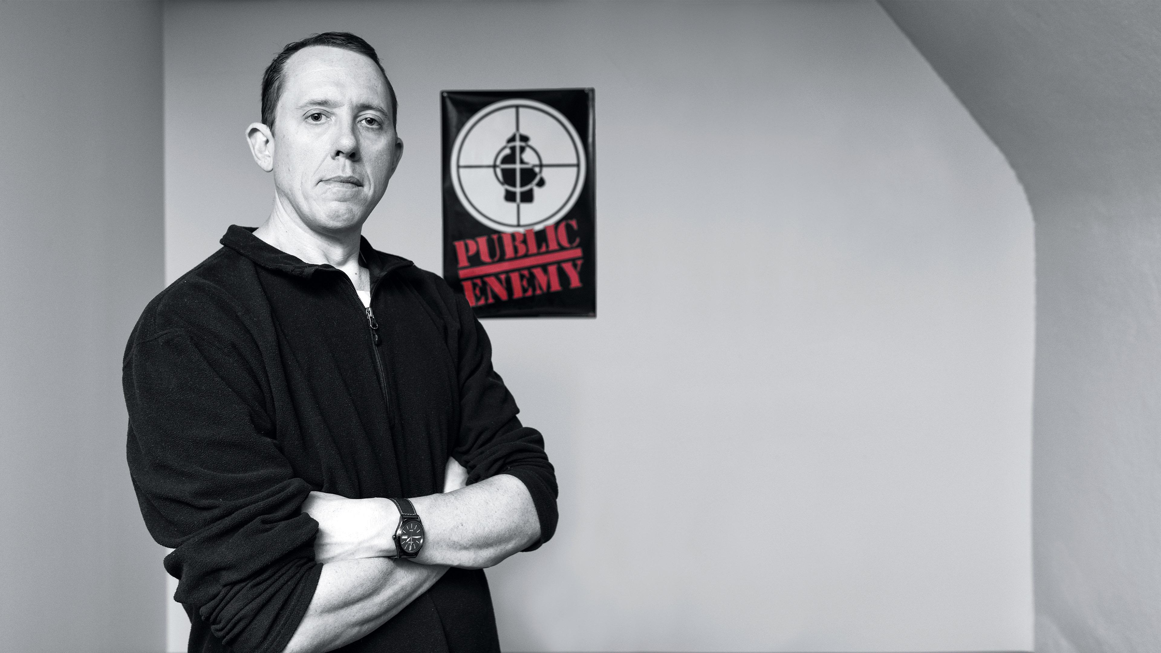 Black and white image of man with his arms crossed in front of a Public Enemy poster 