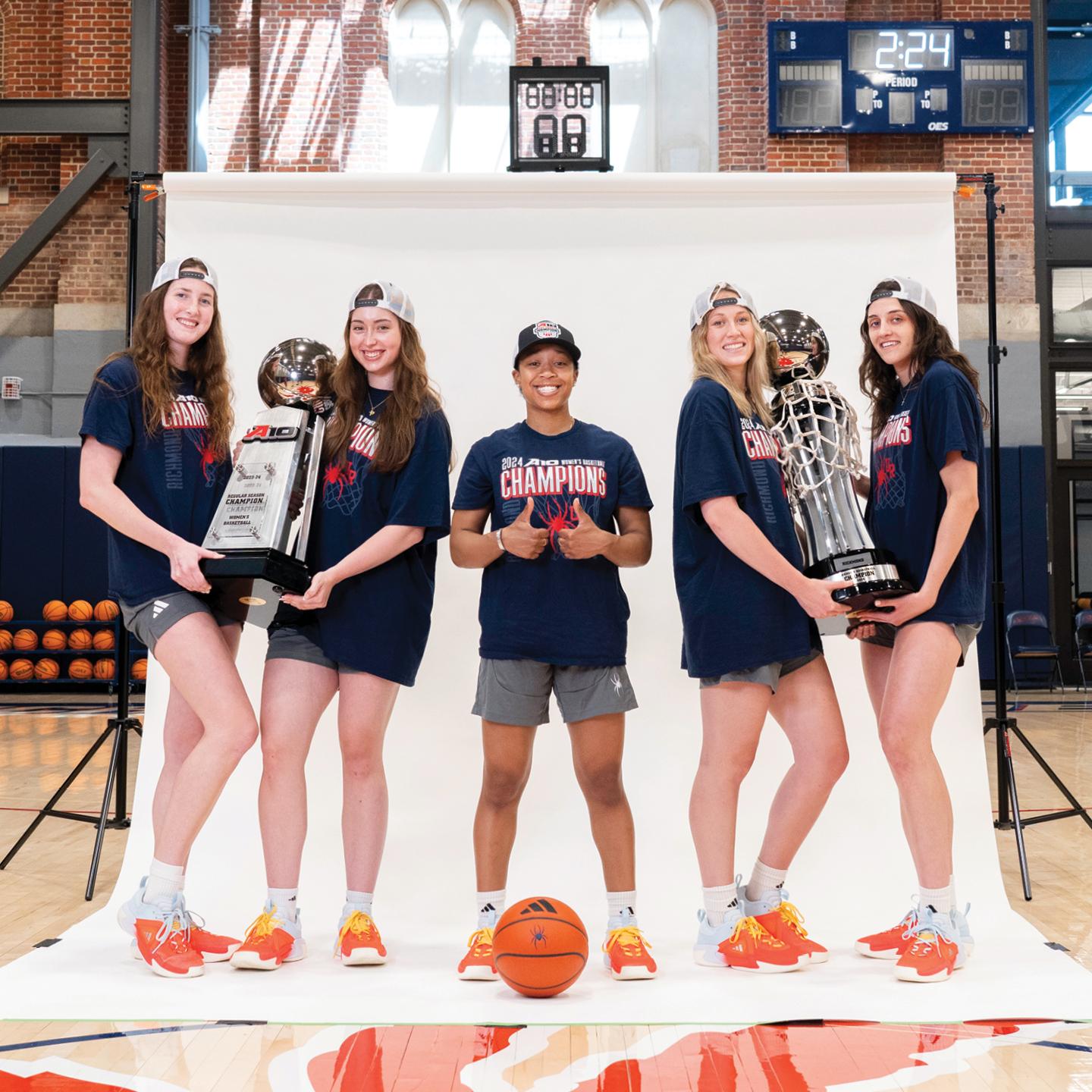 A photograph of five members of the UR Womens' Basketball team holding the A-10 tournament and and A-10 regular season trophies.