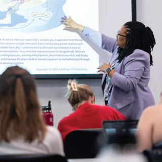 Shannon Jones stands in front of a screen displaying a diagram, presenting to a class of students