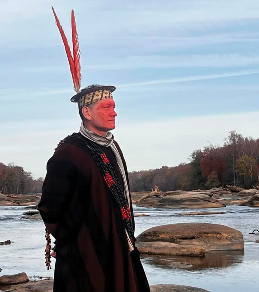 A photo of Wewito Piyãko, leader of a Brazilian Amazonian community, at the James River. He is wearing a black hat with two red feathers and has red paint on his face.