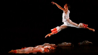 A dancer in red leaps across other dancers laying on the ground.