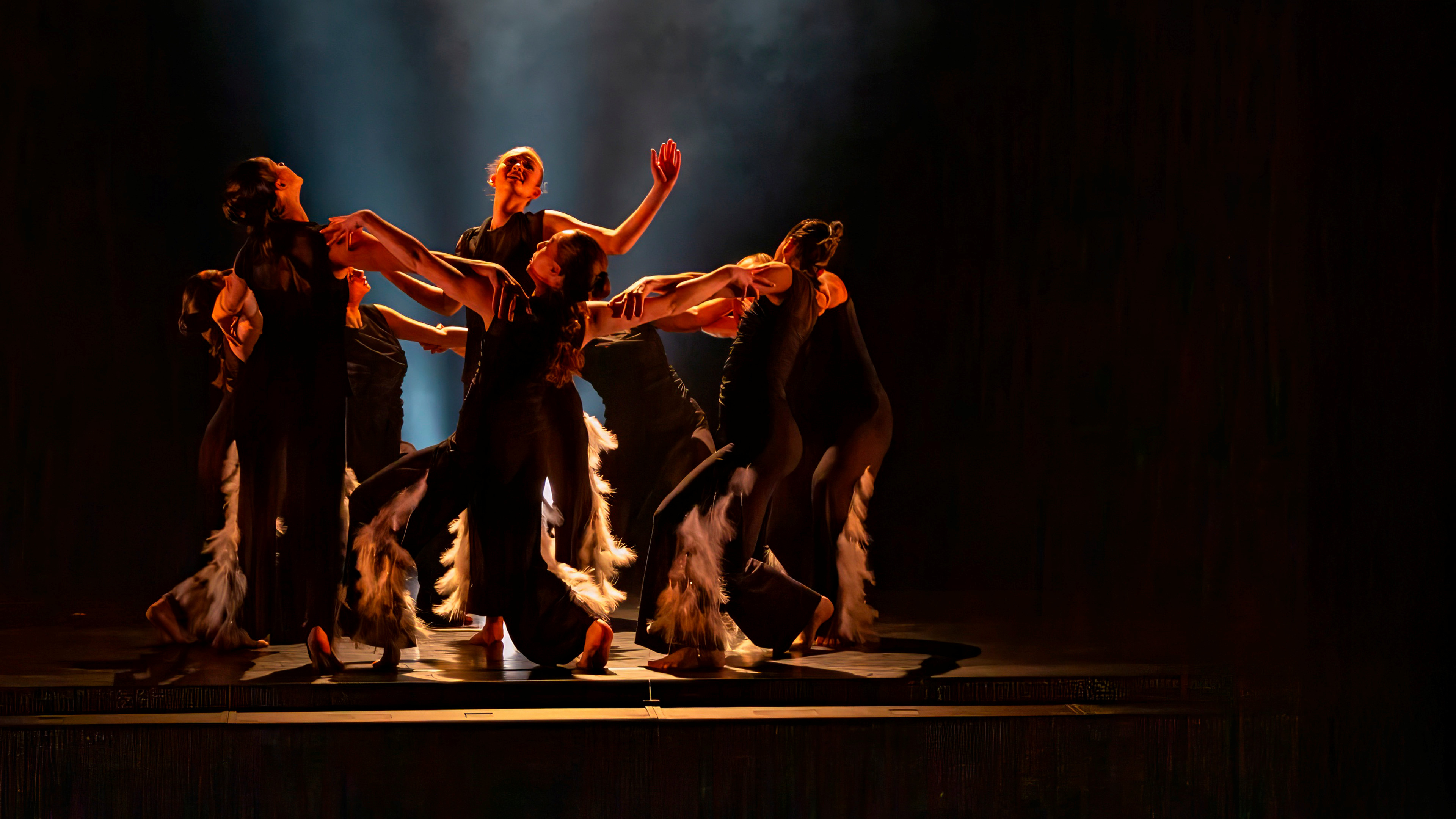 A group of dancers in all black on stage dramatically lit. 