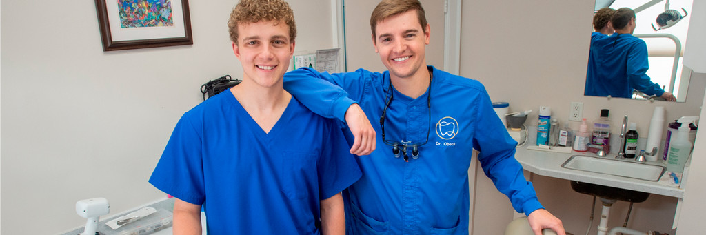 A photo of Jack Bricklemeyer and Dr. John Obeck in Obeck's dental office in Tampa, FL. Both are wearing blue scrubs and smiling at the camera.
