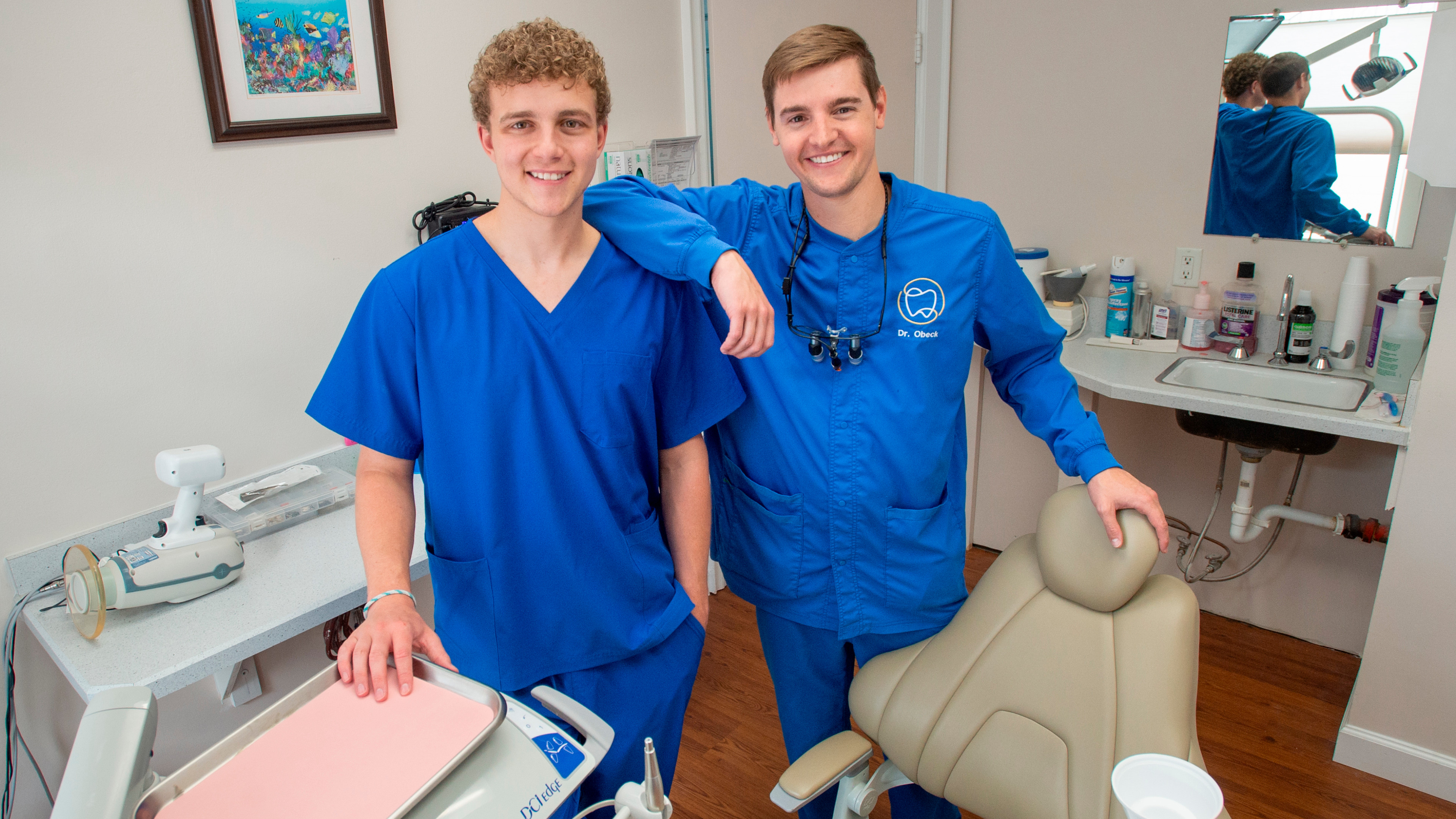 A photo of Jack Bricklemeyer and Dr. John Obeck in Obeck's dental office in Tampa, FL. Both are wearing blue scrubs and smiling at the camera.