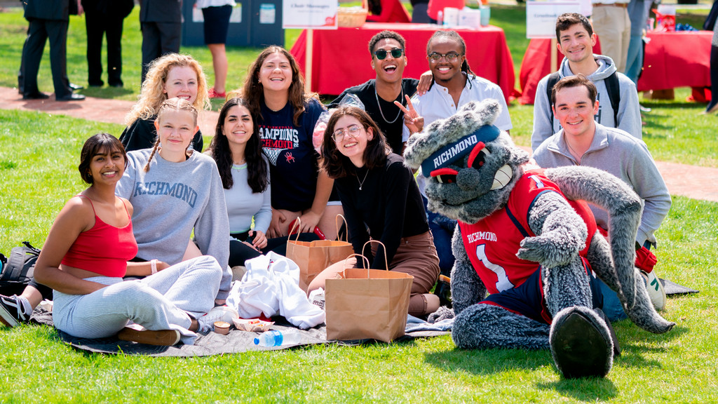 A large group of students, sitting on the grass at Westhampton Green. They are joined by WebstUR and are all smiling into the camera.