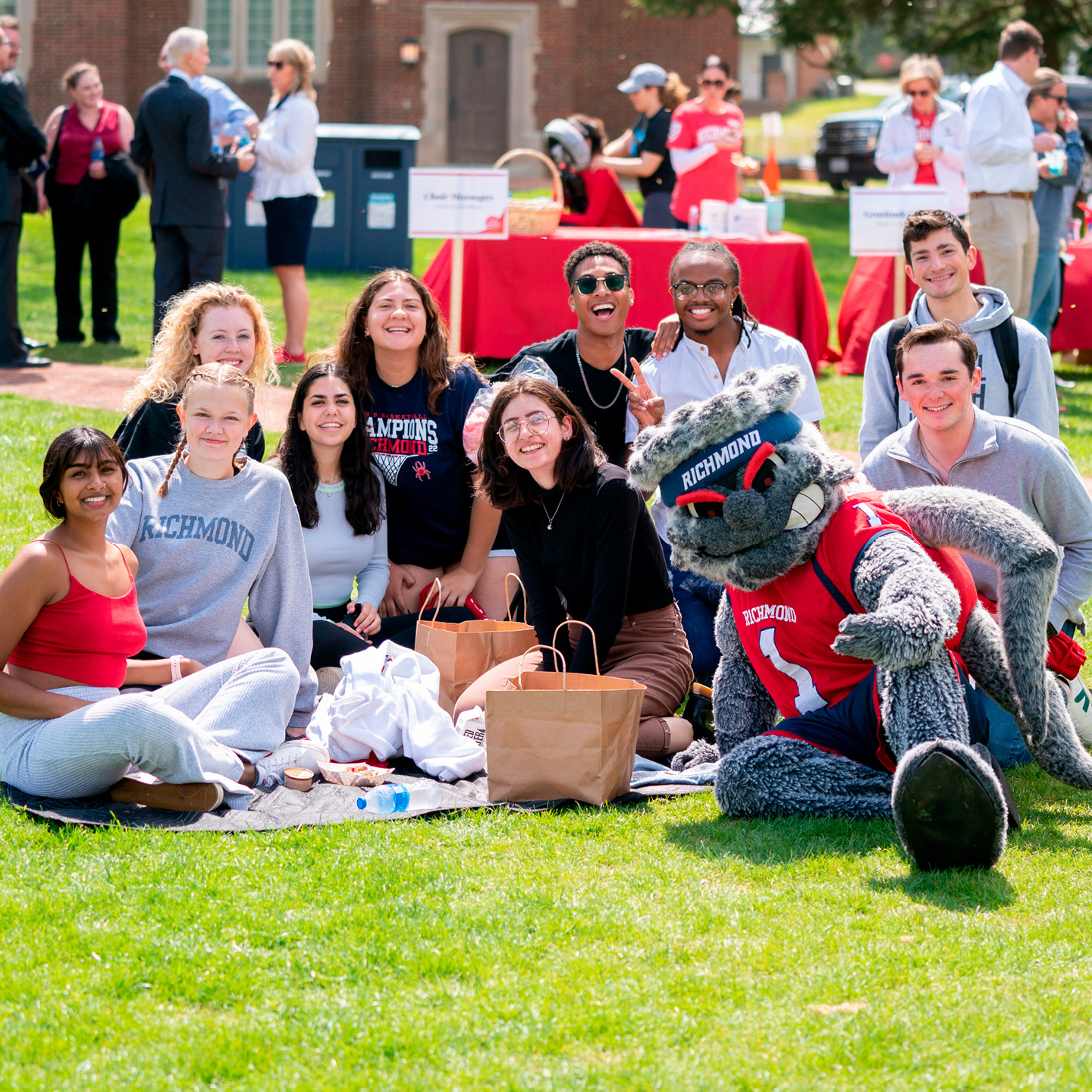 A large group of students, sitting on the grass at Westhampton Green. They are joined by WebstUR and are all smiling into the camera.