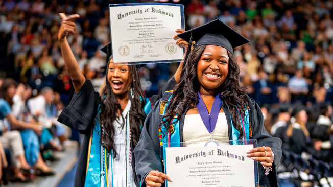 Two students in graduation gowns and mortarboards holding their diplomas up to the camera. The student in theback is holding hers with one hand and pointing to it with the other.