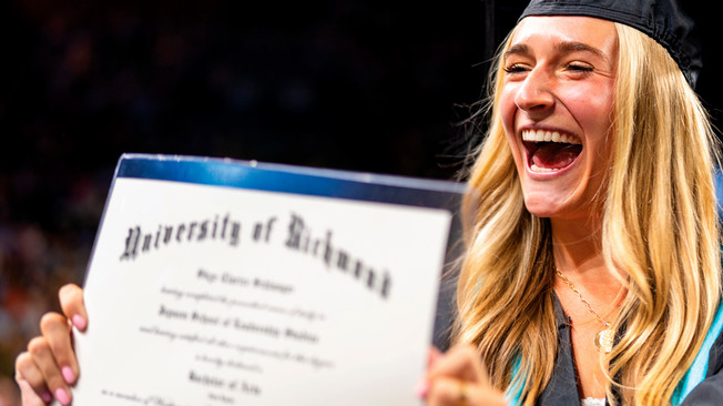A blonde student grinning off camera and holding up her diploma. She is wearing a mortarboard and graduation gown.