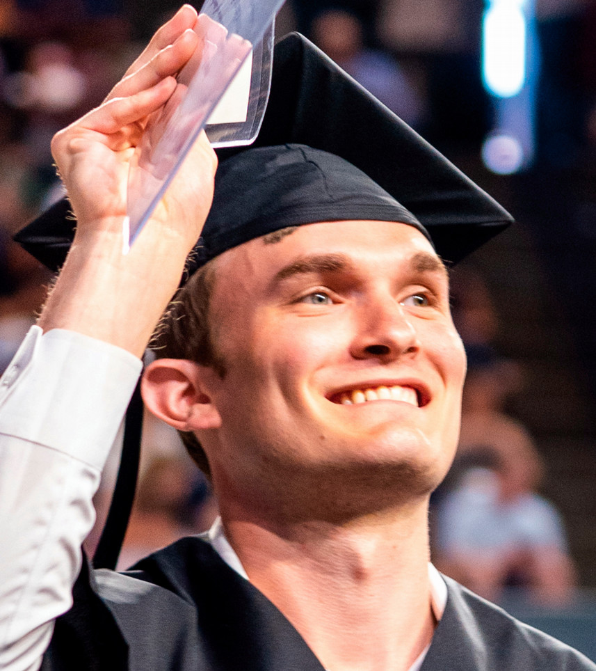 A student smiling looking off camera at someone in the audience. He is wearing a graduation mortarboard and gown.