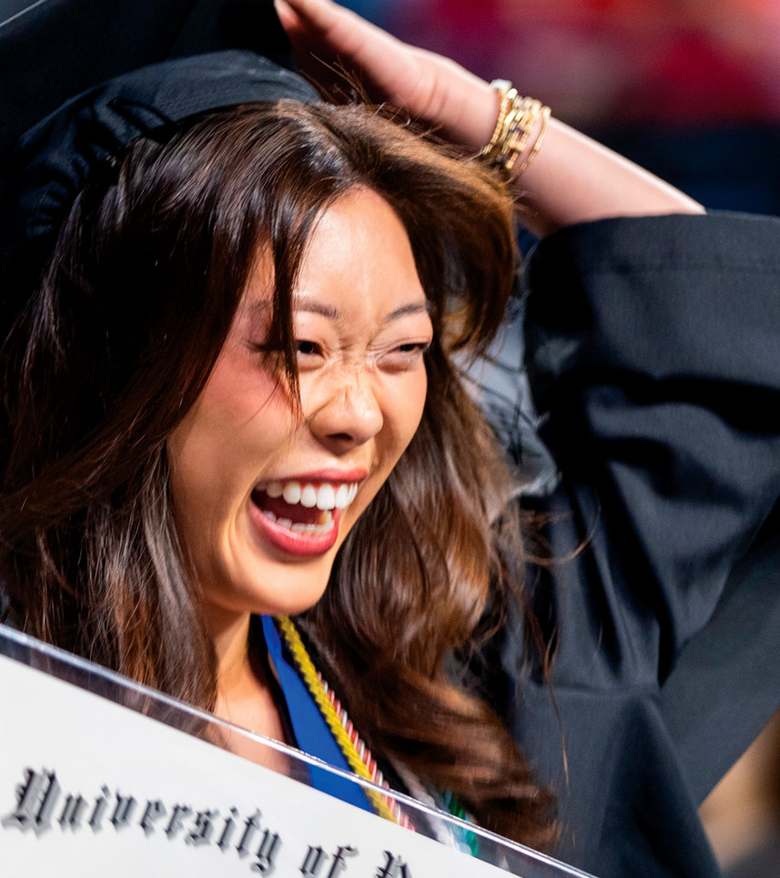 A student smiling holding her diplma. She is wearing a graduation mortarboard and gown, and using one hand to hold her mortarboard on..
