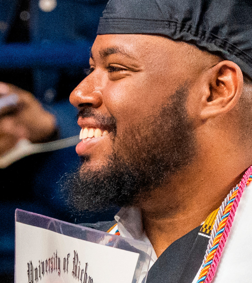 A student in profile, smiling off camera. He is wearing a graduation mortarboard and gown