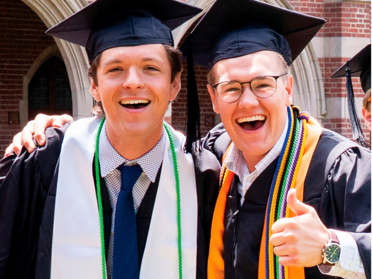 Two students smiling at the camera. They are wearing graduation mortarboards and gowns, and one of them is giving a thumbs up..