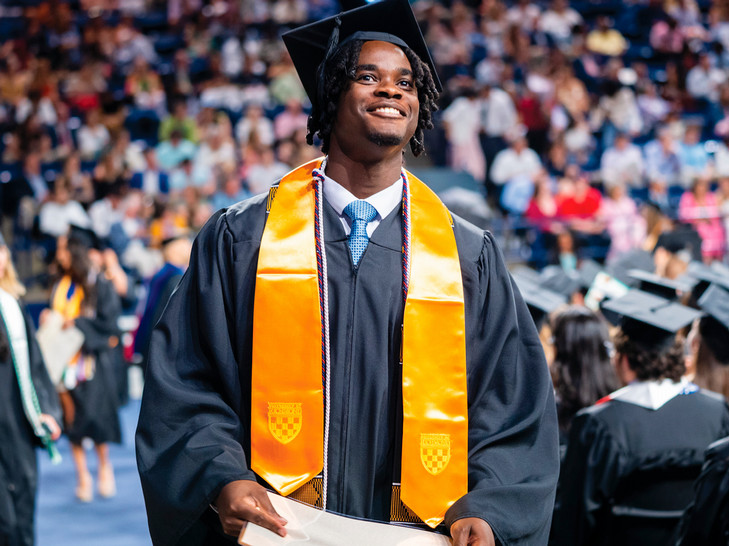 A student smiling off camera, looking up into the stands. He is wearing a graduation mortarboard and gown, and holding his diploma.