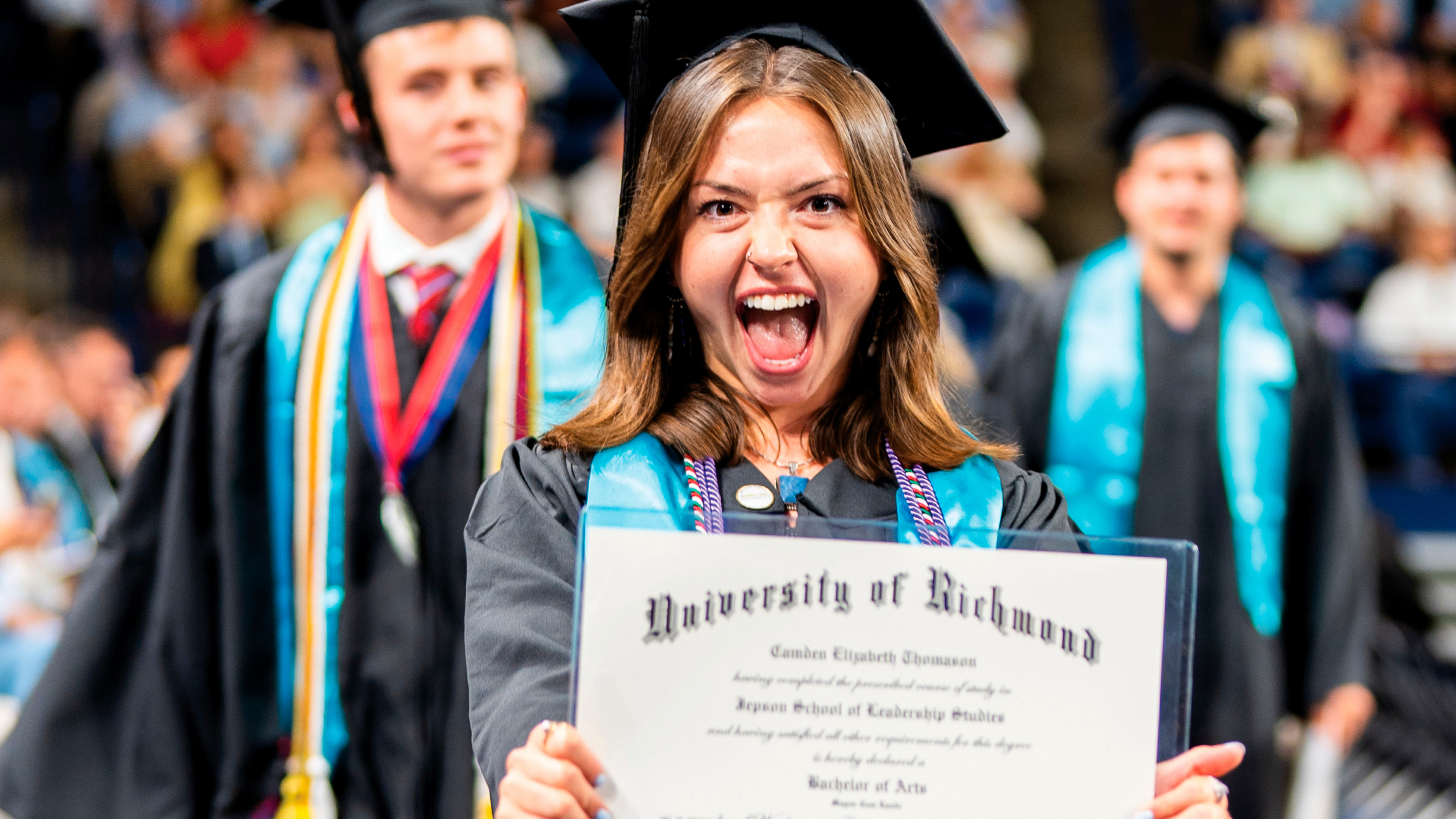 A student smiling with her mouth open. She is wearing a graduation mortarboard and gown, and showing her diploma to the camera.