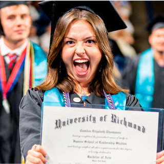 A student smiling with her mouth open. She is wearing a graduation mortarboard and gown, and showing her diploma to the camera.