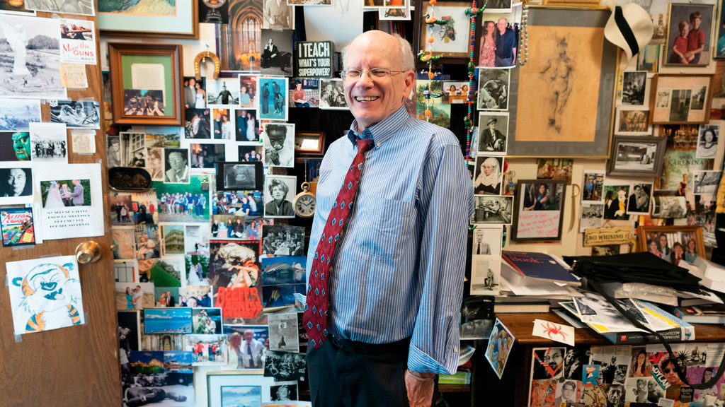 A photo of professor Joe Hoyle. He is smiling at the camera and standing in his office, which is plastered with posters, postcards, art, and other mementos.