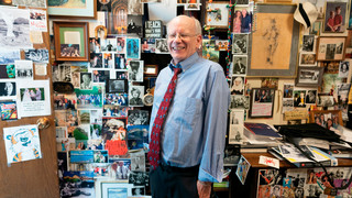 A photo of professor Joe Hoyle. He is smiling at the camera and standing in his office, which is plastered with posters, postcards, art, and other mementos.