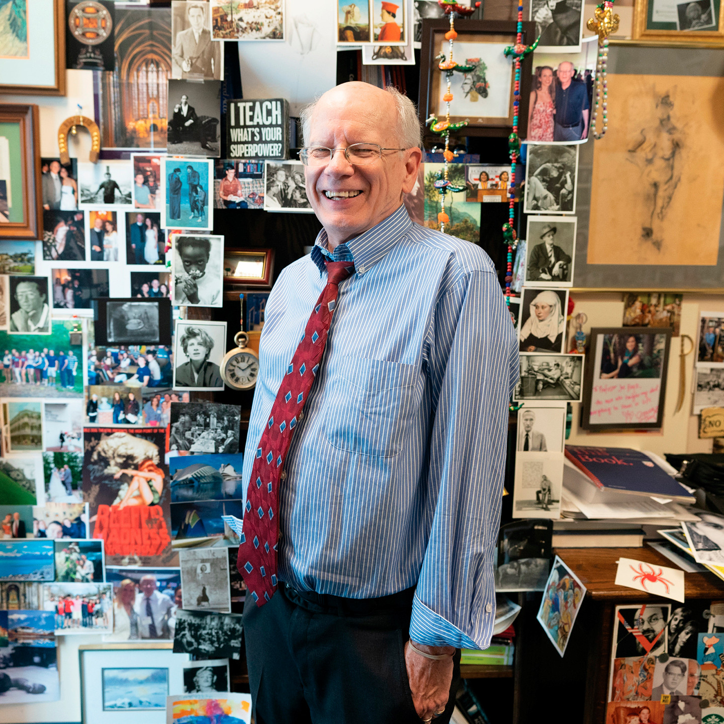 A photo of professor Joe Hoyle. He is smiling at the camera and standing in his office, which is plastered with posters, postcards, art, and other mementos.