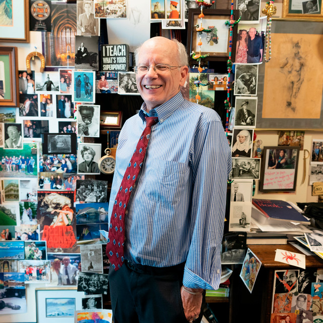 A photo of professor Joe Hoyle. He is smiling at the camera and standing in his office, which is plastered with posters, postcards, art, and other mementos.