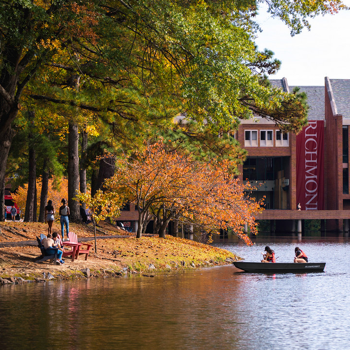 A photo of two students conducting water testing from a boat on Westhampton Lake. They are wearing life jackets and lowering a sample bottle into the water. The commons is visible behind them, and the trees have fall leaves. Other students are visible sitting in Adirondack chairs on the shore of the lake.