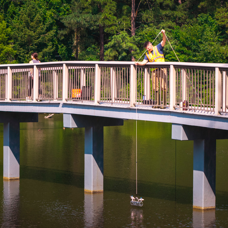 A photo of professor Jonathan Richardson standing on the bridge to the gazebo on Westhampton Lake. He is lowering a device designed to sample water at different depths into the lake.