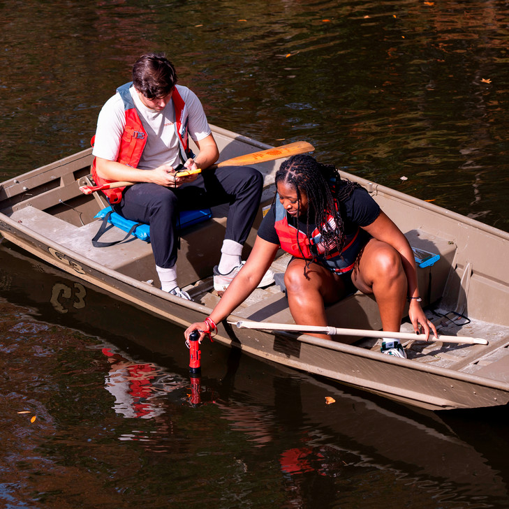 A photo of two students conducting water testing from a boat on Westhampton Lake. They are wearing life jackets and lowering a testing probe into the water.