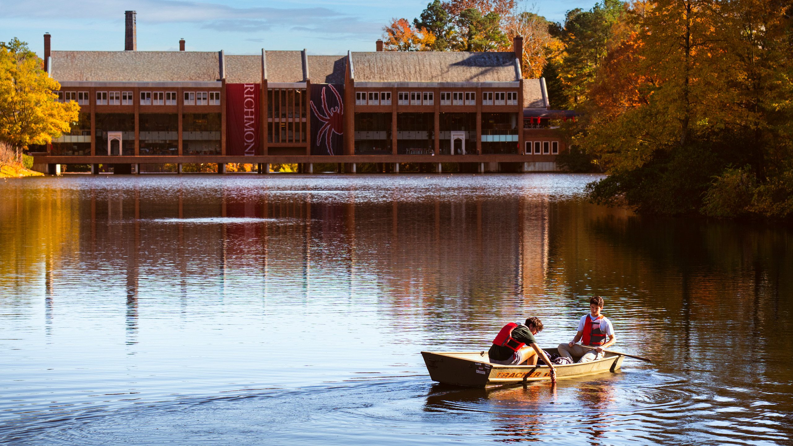 A photo of two students conducting water testing from a boat on Westhampton Lake. They are wearing life jackets and lowering a sample bottle into the water. The commons is visible behind them, and the trees have fall leaves.