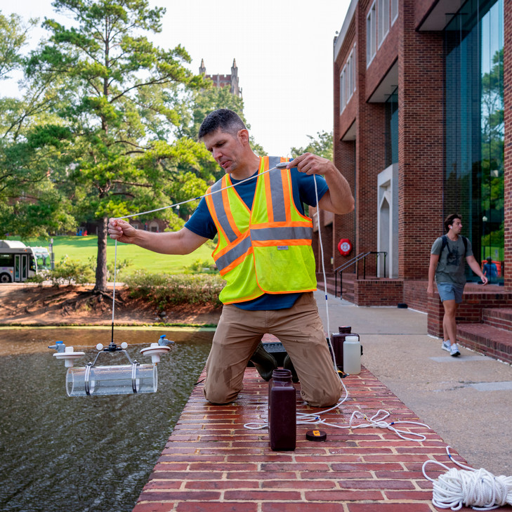 A photo of professor Jonathan Richardson kneeling on the brick walkway near Tyler Haynes Commons, over Westhampton Lake. He is lowering a device designed to sample water at different depths into the lake.