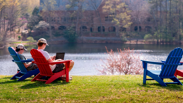 Two students sit in Adirondack chairs by the lake working on laptops. 