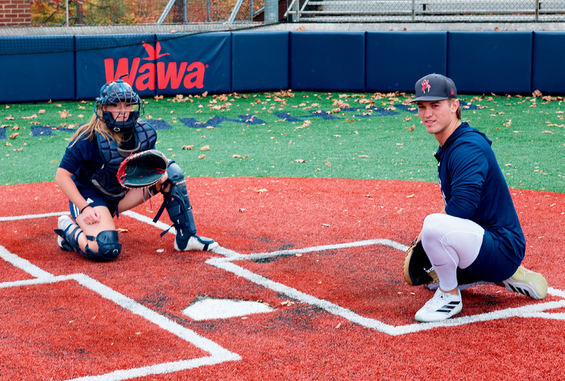 Marisa Snee squats at home plate in baseball catcher's gear, as catcher Dylan Winebrenner gives pointers.