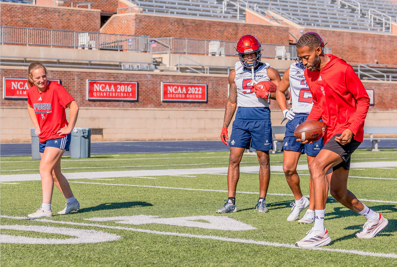 Marisa Snee looks over at spider football wide receivers coach Jerry Taylor as he explains how to run a passing drill as player Ja'vion Griffin looks on.