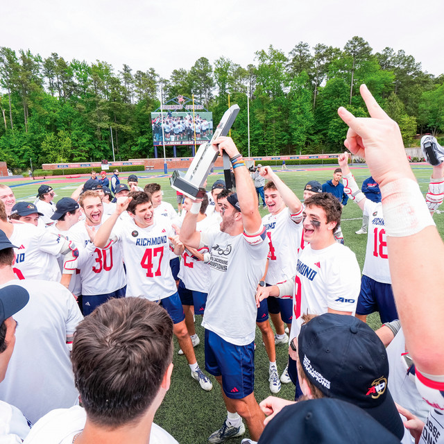 The men's lacrosse team celebrates by hoisting the A-10 trophy above their heads.