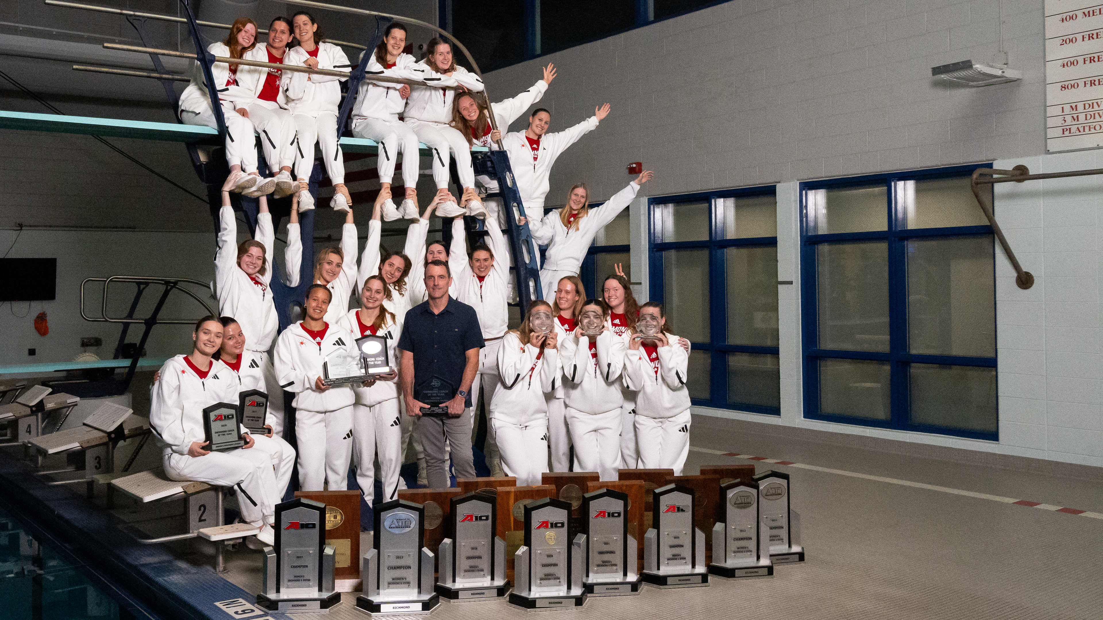 The University of Richmond swim and dive team with coach Matt Barany. They are posing poolside, some of them stacked up on the diving board and stairs. A huge collection of team trophies from over the years sit at thier feet, with smaller trophies being held by team members. The swimmers and divers are wearing white warm up suits.