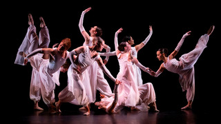 A group of dancers in white dresses on a black backdrop.