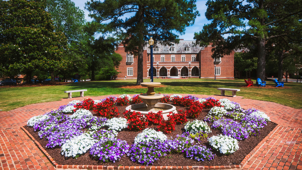 A multi-tiered stone fountain surrounded by a large, circular bed of red, white, and purple flowers on a brick courtyard. In the background, a large brick university building sits behind a green lawn with tall trees.