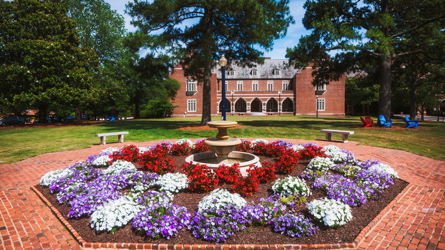 A multi-tiered stone fountain surrounded by a large, circular bed of red, white, and purple flowers on a brick courtyard. In the background, a large brick university building sits behind a green lawn with tall trees.
