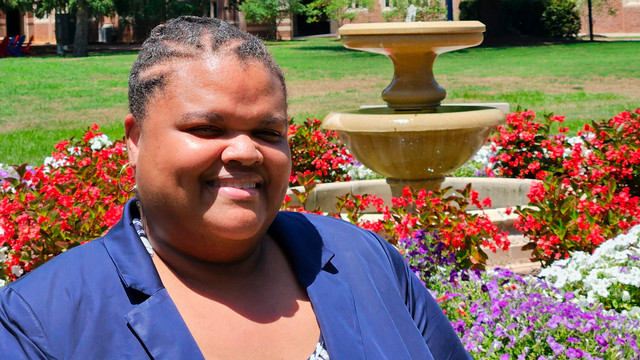 Karle Wells headshot in bright sunshine in front of a fountain and red and white flowers