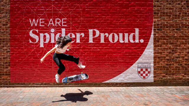 A female skateboarder performs a kickflip in front of a brick mural that reads, "We are Spider Proud."