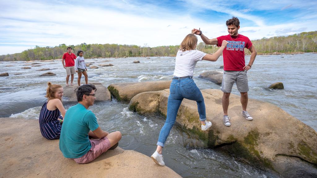 A male student helps a female student cross over large rocks in the James River while other students sit on rocks nearby.