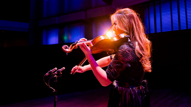 A UR student performs solo violin on the stage of Camp Concert Hall with dramatic back-lighting