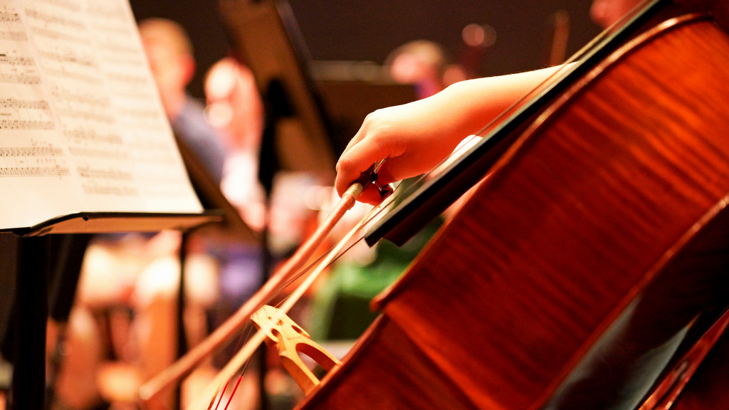 a student's hand bowing a cello during an orchestra rehearsal on stage
